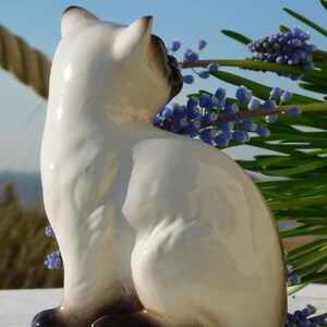 May include: A white ceramic figurine of a Siamese cat sitting with its tail curled around its body. The cat is looking to the right and has black markings on its face and paws. The figurine is set against a background of blue flowers.
