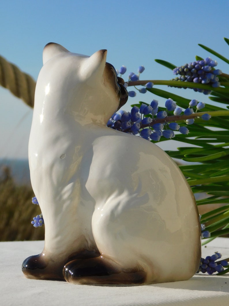 May include: A white ceramic figurine of a Siamese cat sitting with its tail curled around its body. The cat is looking to the right and has black markings on its face and paws. The figurine is set against a background of blue flowers.
