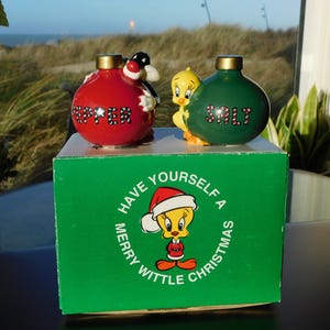 Puede incluir: Un juego de salero y pimentero con temática navideña con personajes de dibujos animados. El pimentero rojo tiene un pingüino, y el salero verde tiene a Piolín. El juego está sobre una caja verde con Piolín con un gorro de Papá Noel y el texto "Have Yourself a Merry Wittle Christmas."