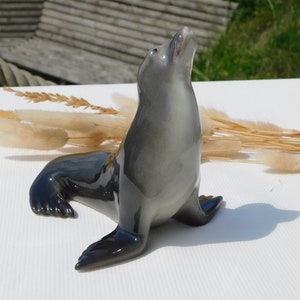 Op de afbeelding: Een grijs keramiek zeehondbeeldje met een glanzende afwerking. De zeehond zit op zijn achterpoten met zijn kop naar achteren gekanteld en zijn mond lichtjes open. Het beeldje staat op een wit oppervlak.