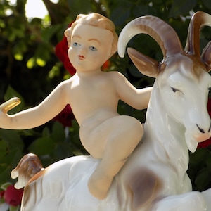 Puede incluir: Una figurita de porcelana de un querubín montando una cabra blanca con marcas marrones. El querubín sostiene un pequeño objeto dorado en su mano. La figurita está colocada sobre un fondo de follaje verde y rosas rojas.