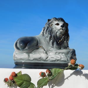 Puede incluir: Figura de león de cerámica en tonos grises y negros, apoyada sobre una base rectangular. El león tiene una melena detallada y se encuentra sobre un cielo azul brillante. Una ramita de bayas con hojas verdes está en primer plano.