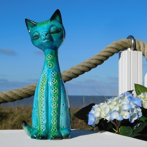 Può includere: Statuetta di gatto in ceramica turchese e verde, con collo lungo e motivi decorativi. Il gatto ha gli occhi chiusi. Lo sfondo comprende un cielo blu, una corda e fiori blu e bianchi.