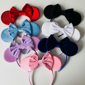 May include: A collection of colorful mouse ear headbands. The headbands feature large, round ears and a matching bow. Colors include red, black, white, blue, purple, and pink. The headbands are made of a soft, velvet-like material.