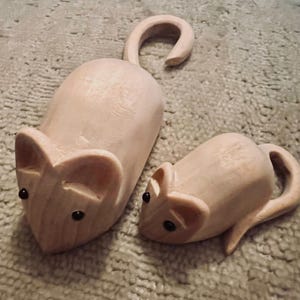 May include: Two wooden mouse figurines. The mice are light brown with black eyes and curved tails. The larger mouse is approximately 13 cm long, and the smaller mouse is about 8 cm long. The figurines are on a textured, light brown surface.