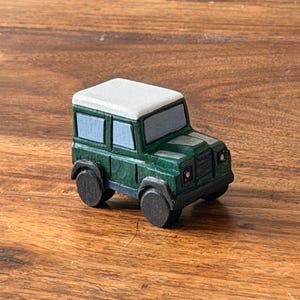 Puede incluir: Un pequeño vehículo de juguete de madera tallado a mano, pintado en verde oscuro con techo blanco, ruedas negras y ventanas azul claro. El juguete tipo SUV presenta un marco cuadrado, una rejilla frontal y textura de madera visible.