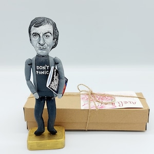 May include: A hand-painted figurine of a man with gray hair and a gray shirt that says "Don't Panic". He is holding a book titled "The Hitchhiker's Guide to the Galaxy". The figurine is standing on a gold base and is in front of a brown gift box with a pink and white ribbon and a tag that says "To you".