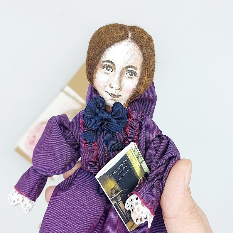May include: A purple doll with brown hair and a white face is holding a book titled "Charlotte Bronte Jane Eyre". The doll is wearing a purple dress with a white lace trim and a black bow.