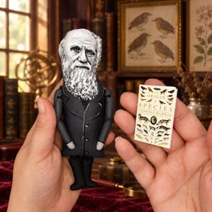 May include: A fabric doll depicting Charles Darwin with a white beard and black coat, held alongside a miniature book titled On the Origin of Species by Charles Darwin, featuring various animal illustrations on its white cover.