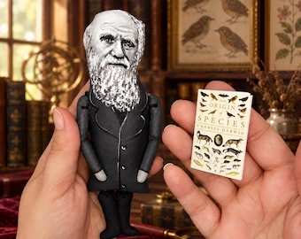 Puede incluir: Una muñeca de tela que representa a Charles Darwin con barba blanca y abrigo negro, sostenida junto a un libro en miniatura titulado On the Origin of Species de Charles Darwin, con varias ilustraciones de animales en su portada blanca.