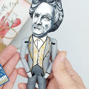 May include: A hand-painted doll of a man with gray hair and a mustache, wearing a gray suit with a gold vest. The doll is holding its hands in front of its waist.