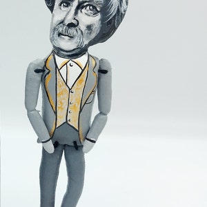 May include: A hand-painted wooden figurine of a man with gray hair and a mustache, wearing a gray suit with a gold vest. The figurine is standing on a gold base.