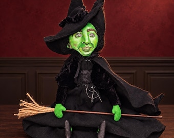 May include: A green-skinned witch doll wearing a black pointed hat, a black velvet-textured jacket, and a long black skirt. The doll has a painted face with visible teeth and holds a small broomstick made of a wooden handle and tan bristles.