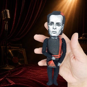 Könnte beinhalten: Eine kleine Stoffpuppe mit einem schwarz-weißen Porträtdruck einer Person mit dunklem Haar und einem roten Akzent. Die Puppe trägt ein schwarzes langärmeliges Outfit mit roten Riemen und hält ein kleines, rotes, dreieckiges Objekt in der Hand.
