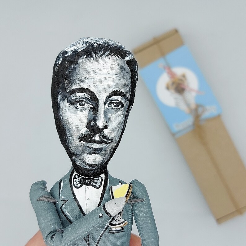 May include: A hand-painted figurine of a man wearing a gray suit and a bow tie. He is holding a small glass in his hand. The figurine is made of wood and has a realistic appearance.