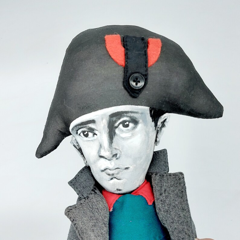 Puede incluir: Una mu&ntilde;eca de tela con un rostro pintado en tonos grises, que lleva un sombrero bicornio negro con un detalle rojo y un peque&ntilde;o bot&oacute;n negro. La mu&ntilde;eca viste un abrigo gris oscuro con cuello rojo y una camisa azul verdoso bajo la chaqueta.