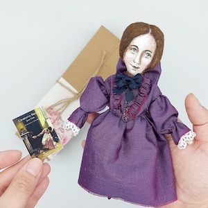 May include: A handmade doll dressed in a purple dress with a ruffled collar and a black bow. The doll has brown hair and painted features. The doll is holding a book titled "Charlotte Bronte Jane Eyre".