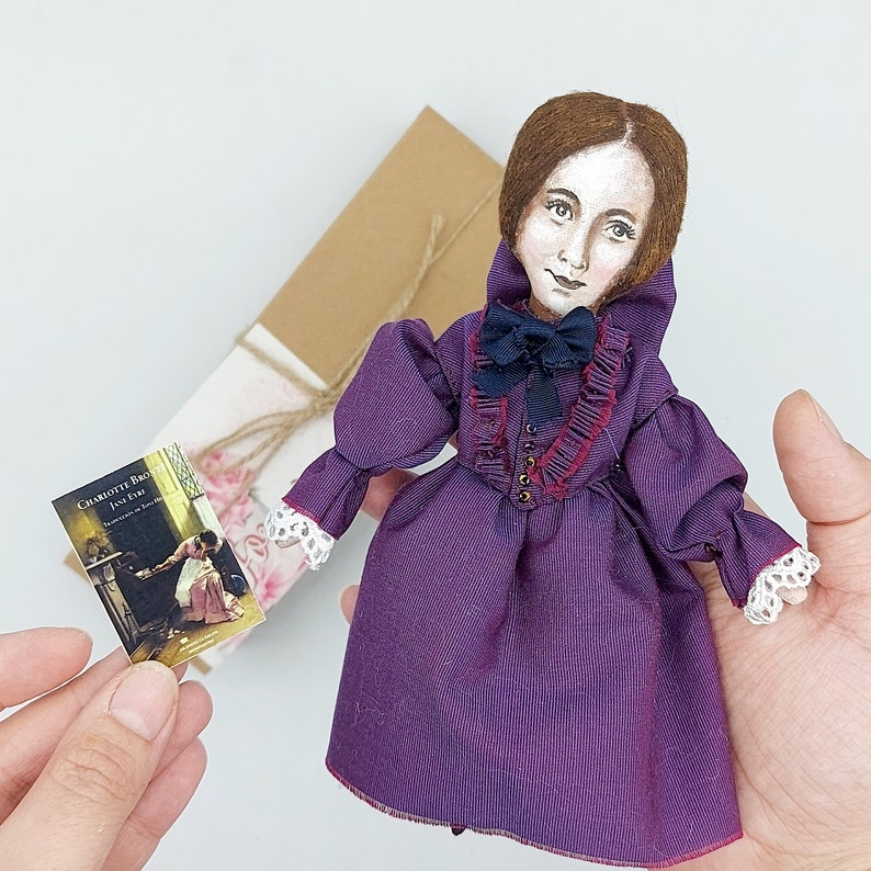 May include: A handmade doll dressed in a purple dress with a ruffled collar and a black bow. The doll has brown hair and painted features. The doll is holding a book titled "Charlotte Bronte Jane Eyre".