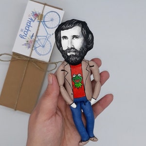 May include: A hand-painted doll with a brown jacket, red shirt, blue jeans, and a green frog graphic. The doll has a black beard and brown hair. The doll is holding a small card that says "Be Happy" with a bicycle graphic.