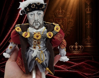 Puede incluir: Una muñeca de tela que representa al rey Enrique VIII, con un sombrero de terciopelo negro con plumas blancas, una túnica roja oscura con adornos dorados y un jubón gris. La figura lleva un collar de tono dorado con detalles rojos y una espada.