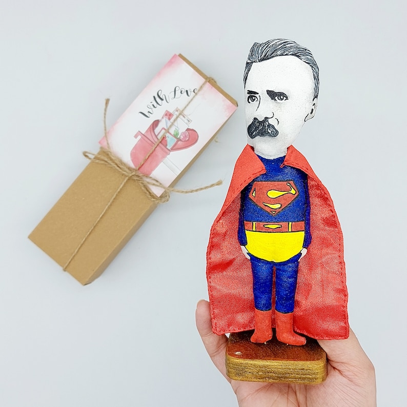 Puede incluir: Una figurita pintada a mano de un hombre con una capa roja y un traje azul de Superman. La figurita tiene bigote y una expresi&oacute;n seria. La figurita est&aacute; de pie sobre una base de madera. El fondo incluye una bolsa de regalo marr&oacute;n con una tarjeta rosa y blanca que dice "With Love".