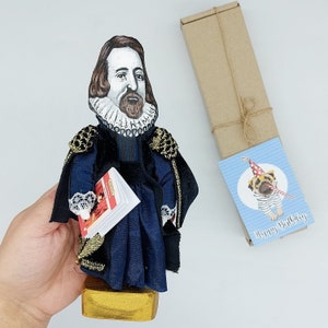 Op de afbeelding: Een handgemaakte pop van een man in een zwart en blauw kostuum met een witte kraag. Hij houdt een klein boek in zijn linkerhand. De pop staat op een houten voet. De pop is een verjaardagscadeau.