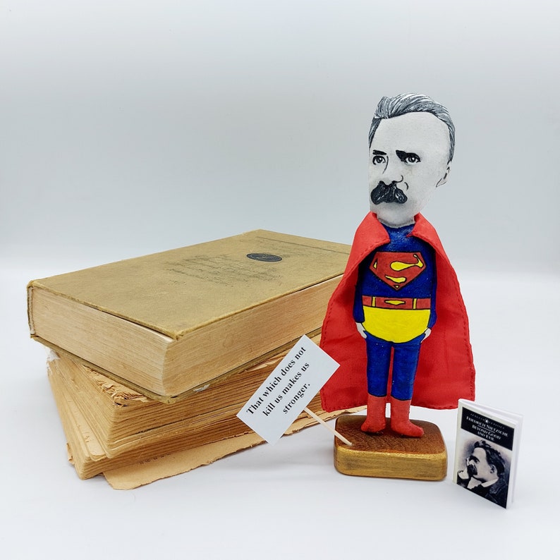 Puede incluir: Una figurita artesanal de un hombre vestido como Superman. El hombre tiene barba y bigote blancos y lleva una capa roja y un traje azul con un cintur&oacute;n amarillo. La figurita est&aacute; de pie sobre una base de madera. La cita "Lo que no nos mata nos hace m&aacute;s fuertes." est&aacute; escrita en un cartel blanco delante de la figurita.