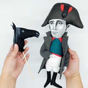 Puede incluir: Una mu&ntilde;eca de tela que representa a una figura hist&oacute;rica con un sombrero bicornio negro con un detalle rojo, abrigo gris, chaleco verde azulado, calzones blancos y botas negras, acompa&ntilde;ada de una marioneta de cabeza de caballo negra.