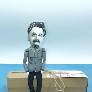 May include: A hand-crafted doll of a man with a mustache and glasses, wearing a gray shirt and black pants. The doll is sitting on a brown box with a white label that says "Love you".