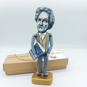 May include: A hand-painted wooden figurine of a man holding a book titled "Adventures of Tom Sawyer". The figurine is wearing a gray suit with a gold vest and is standing on a wooden base.