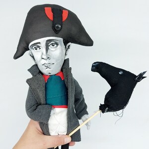Puede incluir: Una mu&ntilde;eca de tela que representa una figura hist&oacute;rica con un sombrero bicornio negro con un detalle rojo y un bot&oacute;n negro, un abrigo gris, un chaleco verde azulado con ribete rojo y pantalones blancos. La mu&ntilde;eca sostiene un caballo negro.