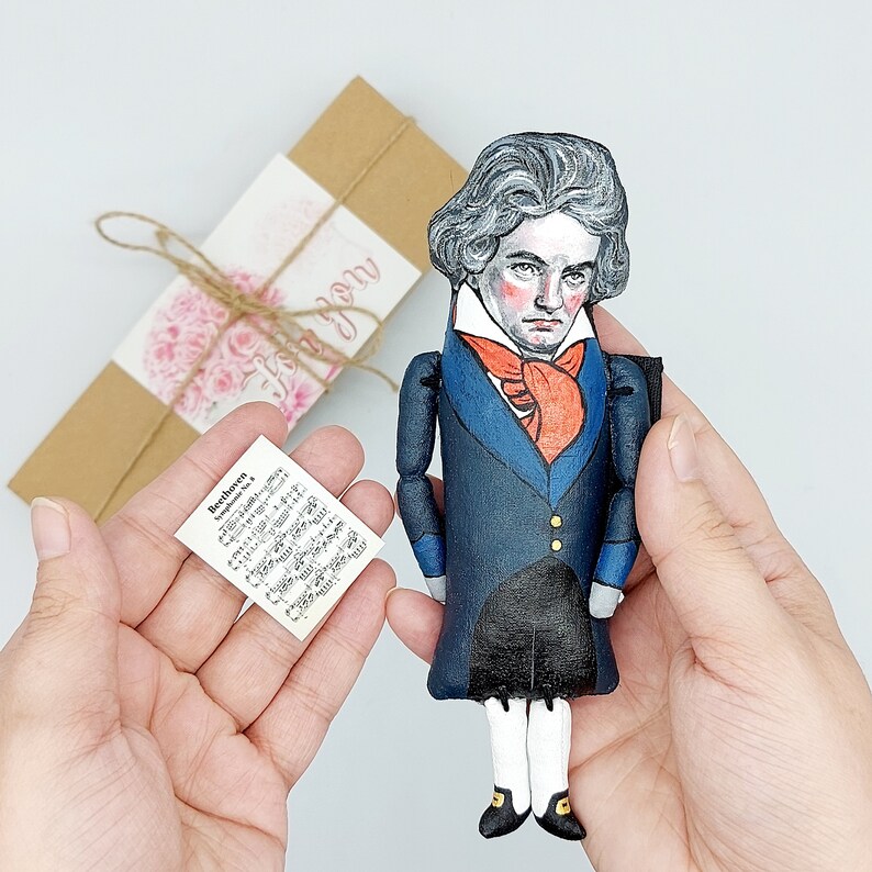 May include: A hand-painted doll of a man wearing a blue suit with a red bow tie. The doll has a white face with painted black hair and brown eyes. The doll is holding a sheet of music with the text "Beethoven Symphony No. 5" printed on it.