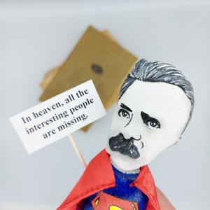 Puede incluir: Una figurita pintada a mano de un hombre con bigote que lleva un traje de superh&eacute;roe rojo y azul. Sostiene un cartel que dice "En el cielo, faltan todas las personas interesantes."