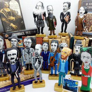 May include: A collection of handmade felt figures of famous authors and artists, including Frida Kahlo, Vincent van Gogh, and Edgar Allan Poe. Each figure is dressed in a unique outfit and holds a prop related to their work. The figures are displayed on a shelf with a variety of books.
