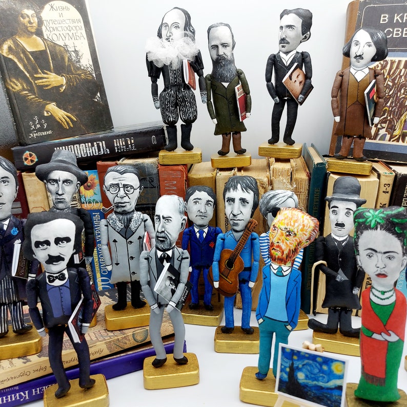 May include: A collection of handmade felt figures of famous authors and artists, including Frida Kahlo, Vincent van Gogh, and Edgar Allan Poe. Each figure is dressed in a unique outfit and holds a prop related to their work. The figures are displayed on a shelf with a variety of books.