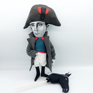 Puede incluir: Una mu&ntilde;eca de tela que representa una figura hist&oacute;rica con un sombrero bicornio gris con un detalle rojo, un abrigo largo gris, un chaleco verde azulado y pantalones blancos. La mu&ntilde;eca tiene la cara pintada y un caballito de palo negro.