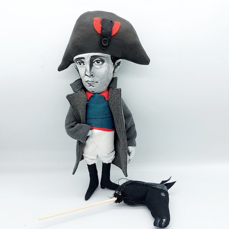 Puede incluir: Una mu&ntilde;eca de tela que representa una figura hist&oacute;rica con un sombrero bicornio gris con un detalle rojo, un abrigo largo gris, un chaleco verde azulado y pantalones blancos. La mu&ntilde;eca tiene la cara pintada y un caballito de palo negro.