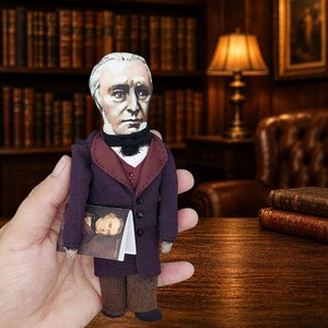 May include: A small fabric doll depicting a historical figure with gray hair, wearing a dark purple coat with a maroon lapel, a white shirt, and a black cravat. The doll holds a miniature book featuring a portrait of the same figure on its cover.