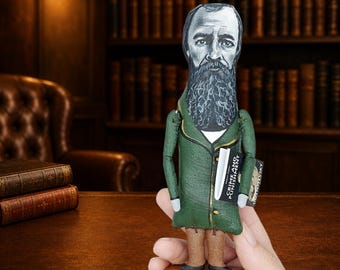 May include: A fabric doll depicting a bearded man with a gray face, wearing a dark green coat with gold trim and brown trousers. The doll holds a miniature book titled Crime and Punishment by Dostoevsky and stands on a rectangular wooden base.