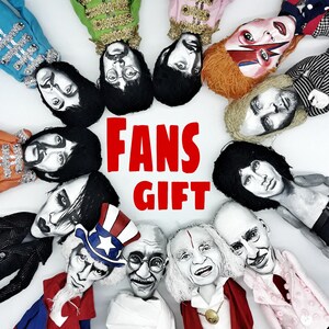 Puede incluir: Una colecci&oacute;n de doce mu&ntilde;ecos pintados a mano dispuestos en c&iacute;rculo, que representan figuras famosas, incluidos m&uacute;sicos e iconos hist&oacute;ricos. El texto FANS GIFT est&aacute; impreso en letras rojas en negrita en el centro del c&iacute;rculo.