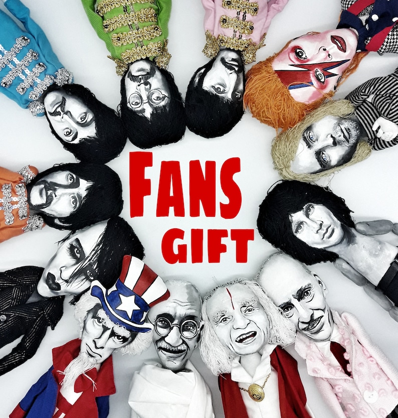 Puede incluir: Una colecci&oacute;n de doce mu&ntilde;ecos pintados a mano dispuestos en c&iacute;rculo, que representan figuras famosas, incluidos m&uacute;sicos e iconos hist&oacute;ricos. El texto FANS GIFT est&aacute; impreso en letras rojas en negrita en el centro del c&iacute;rculo.