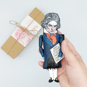 May include: A hand-painted doll of a man wearing a blue jacket with a red bow tie and holding a sheet of music with the text "Beethoven Symphony No. 5".