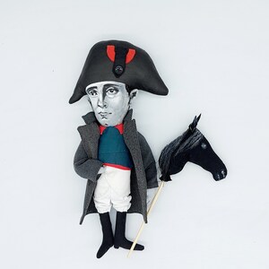 Puede incluir: Una mu&ntilde;eca de tela que representa una figura hist&oacute;rica con un sombrero bicornio negro con un detalle rojo, abrigo gris, chaleco verde azulado, calzones blancos y botas negras. La mu&ntilde;eca sostiene un caballo de juguete negro en un palo.