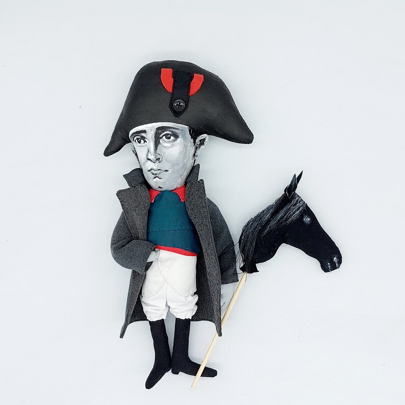 Puede incluir: Una mu&ntilde;eca de tela que representa una figura hist&oacute;rica con un sombrero bicornio negro con un detalle rojo, abrigo gris, chaleco verde azulado, calzones blancos y botas negras. La mu&ntilde;eca sostiene un caballo de juguete negro en un palo.