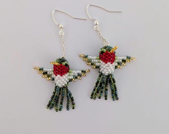 May include: A pair of handmade hummingbird earrings. Each earring features a beaded hummingbird with a red throat, white chest, and green and gold wings. The tail feathers are dark green and blue. The earrings hang from silver-colored hooks.
