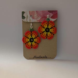 May include: Handmade beaded flower earrings in shades of red, orange, yellow, and black. The earrings are attached to silver-colored hooks and are displayed on a brown card with floral accents. The card has the text "...Handmade..."