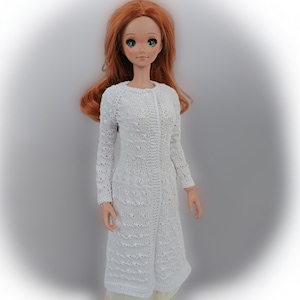 May include: A doll wearing a long, white, knitted coat. The coat has a button-up front and long sleeves. The doll has red hair and blue eyes. The coat appears to be handmade and has a textured pattern.