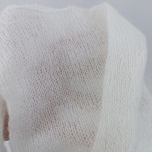 May include: Close-up of a soft, white, sheer fabric. The material is loosely woven, creating a delicate, open texture. The fabric is draped, showing folds and shadows, suggesting a lightweight textile.