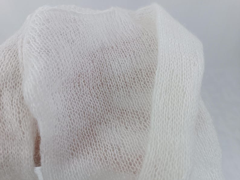 May include: Close-up of a soft, white, sheer fabric. The material is loosely woven, creating a delicate, open texture. The fabric is draped, showing folds and shadows, suggesting a lightweight textile.