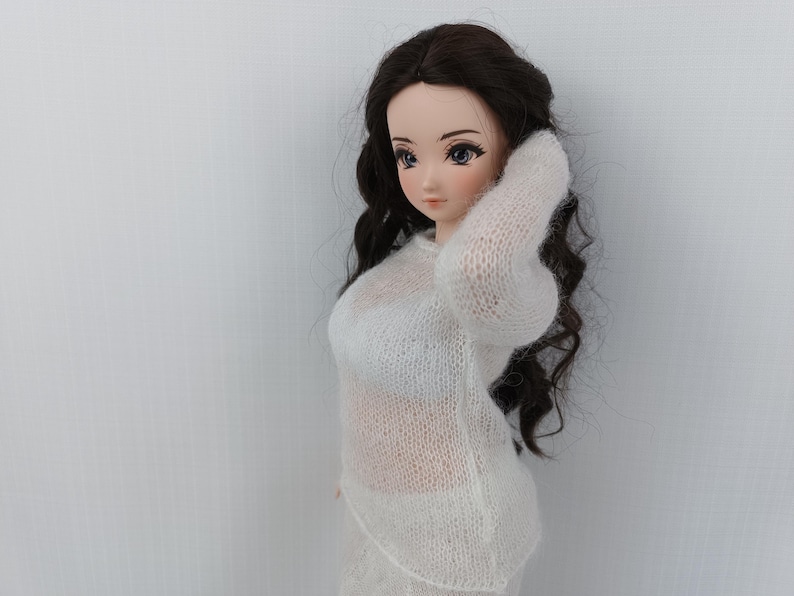 May include: A doll with dark, wavy hair, dressed in a white, sheer knitted sweater and skirt. The doll is posed with an arm raised. The outfit is likely hand-knitted or crocheted. The doll has blue eyes and is set against a plain white backdrop.